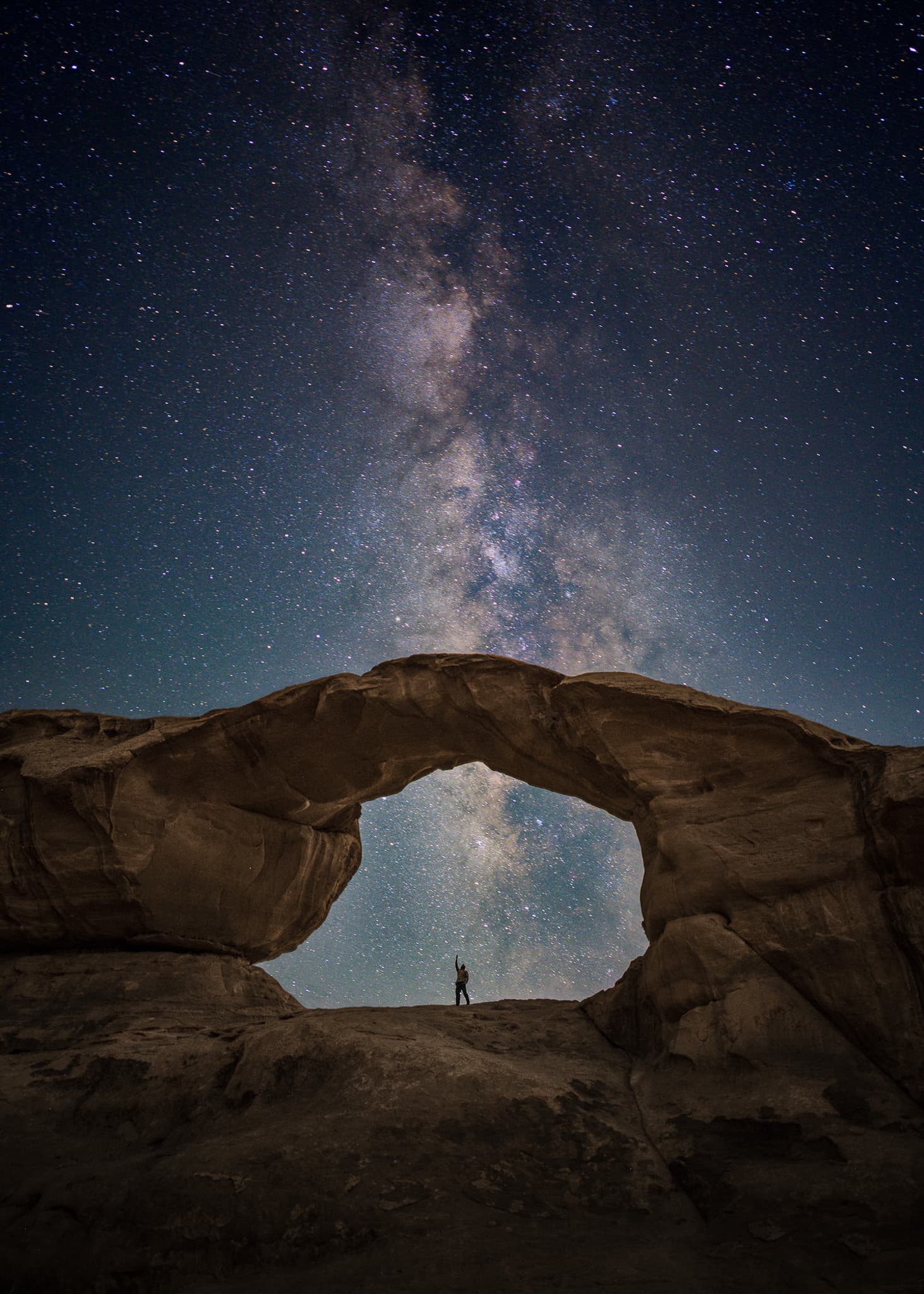 The Milky Way rising behind a natural rock arch with a lone figure beneath it