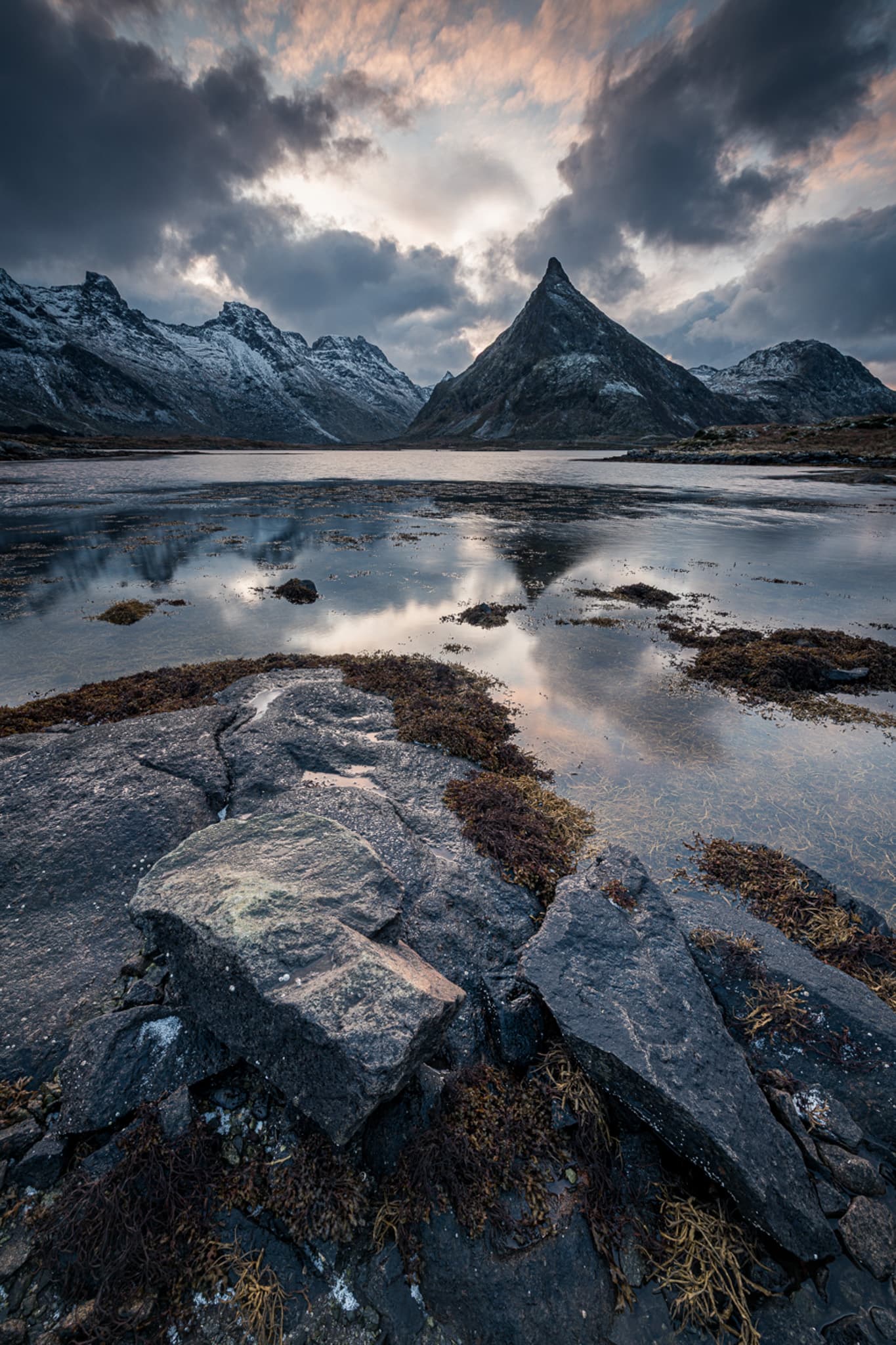 A sharp mountain peak reflected in coastal water beneath dramatic winter clouds